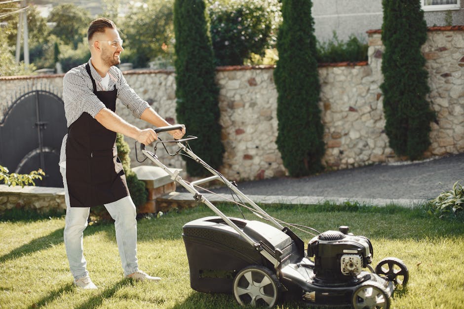 A man wearing a black apron and light-colored trousers is engaged in lawn care work in a well-maintained garden. He is pushing a petrol-powered lawn mower over a lush, green grass lawn, which appears 