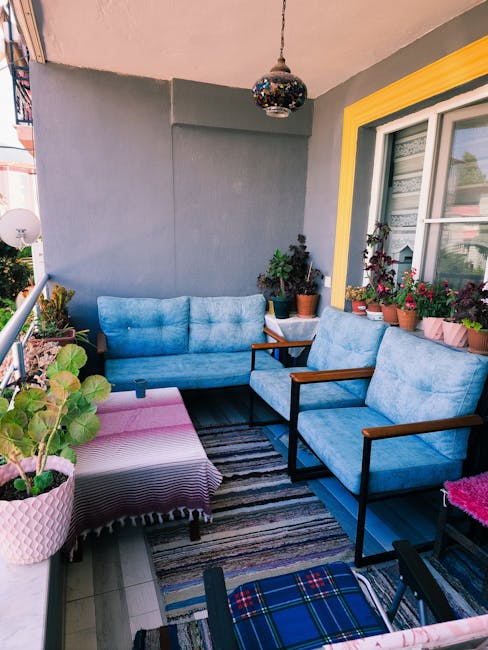 A cozy balcony garden features a trio of blue cushioned chairs with dark wooden armrests arranged around a striped outdoor rug. To the left, a pink ceramic pot with a large leafy green plant sits on a